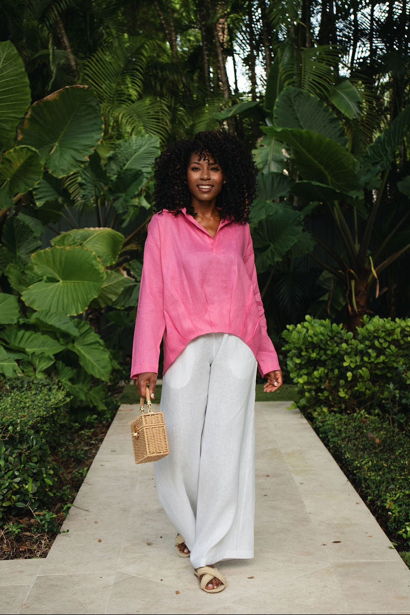A woman walking on a stone path surrounded by lush green tropical plants, wearing a bright pink long-sleeve collared shirt, 100% linen white wide-leg pants, and holding a small beige handbag.