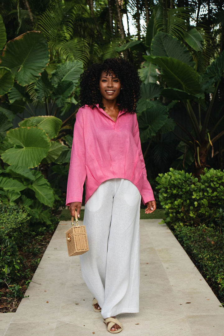 A woman walking on a stone path surrounded by lush green tropical plants, wearing a fuchsia pink long-sleeve linen collared shirt, white wide-leg pants, and holding a small beige handbag.