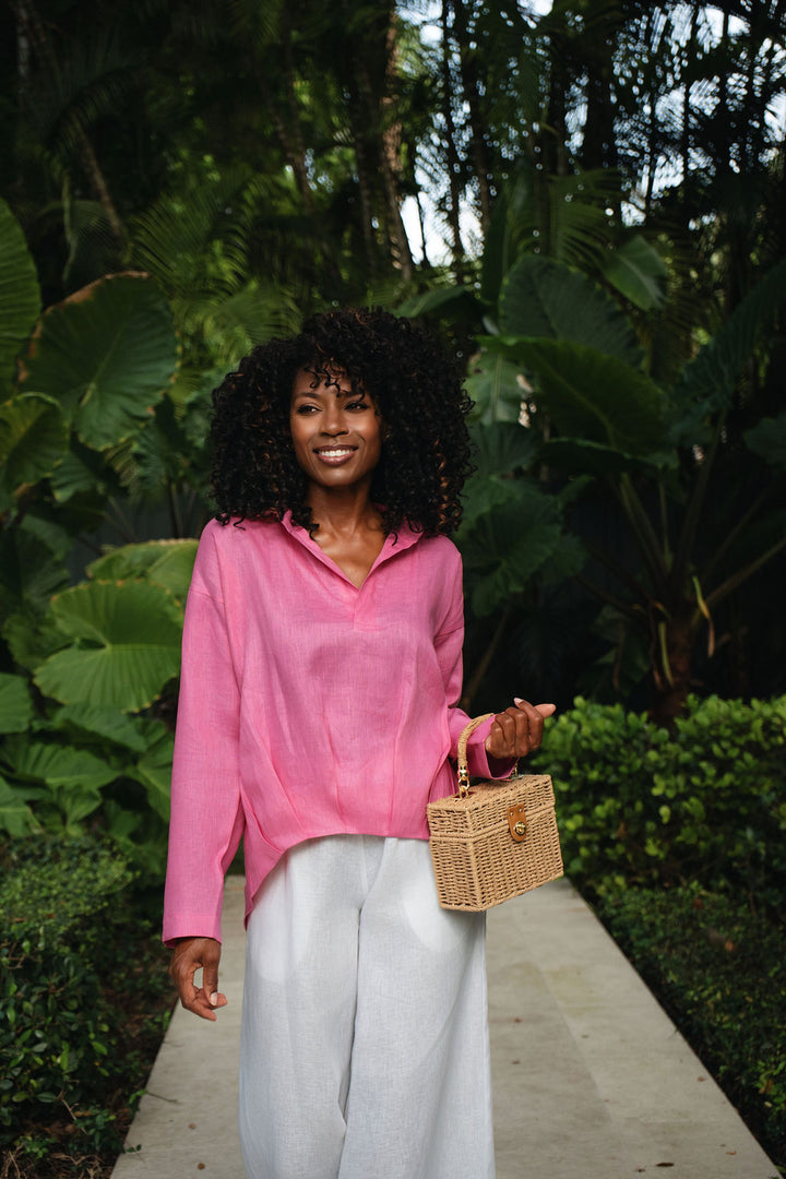 A woman with curly black hair stands on a garden path wearing a bright pink long-sleeve collared linen blouse and white wide-leg pants, holding a small woven tan handbag.