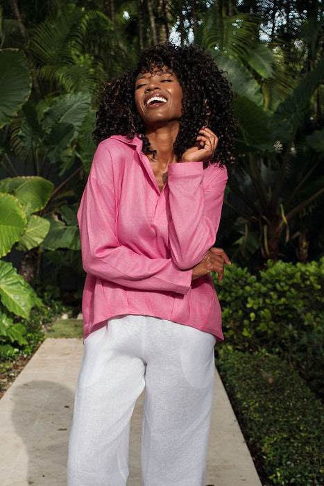 A woman stands on a stone pathway in a lush garden, wearing a bright pink long-sleeve top and white wide-leg pants.