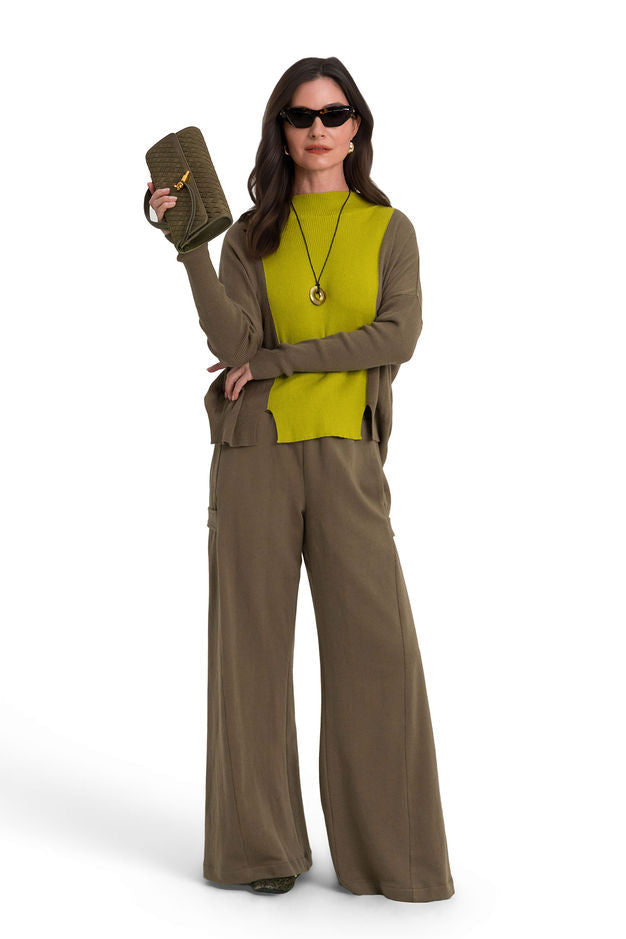 A woman modeling a matching olive green lounge set consisting of a long-sleeve pima cotton sweater and 100% cotton french terry wide-leg pants, styled with a bright yellow-green knit vest, sunglasses, and a clutch bag against a plain white background.