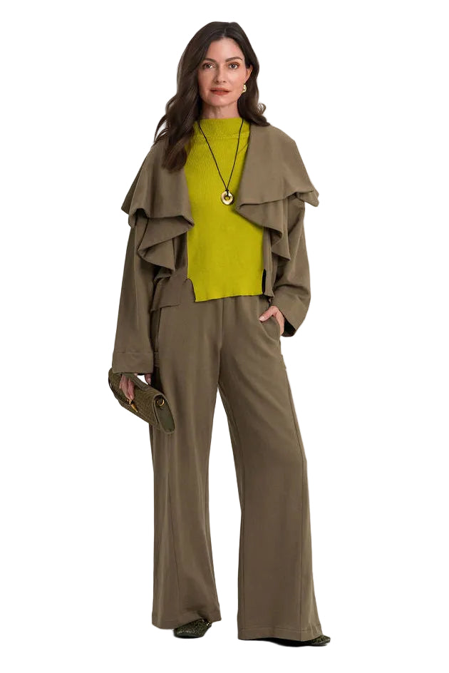 A woman modeling a matching 100% cotton olive green two-piece set consisting of a draped long-sleeve top and wide-leg trousers, styled with a bright yellow-green turtleneck and a gold pendant necklace, against a plain white studio background.