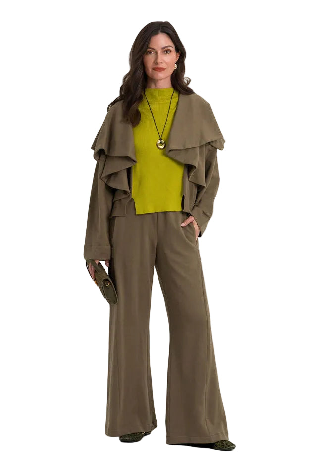 A woman stands against a plain light gray studio background wearing a coordinated outfit consisting of wide-leg olive-brown pants, a bright yellow-green ribbed top, and a draped olive-brown jacket. She is also wearing a long gold-toned necklace.