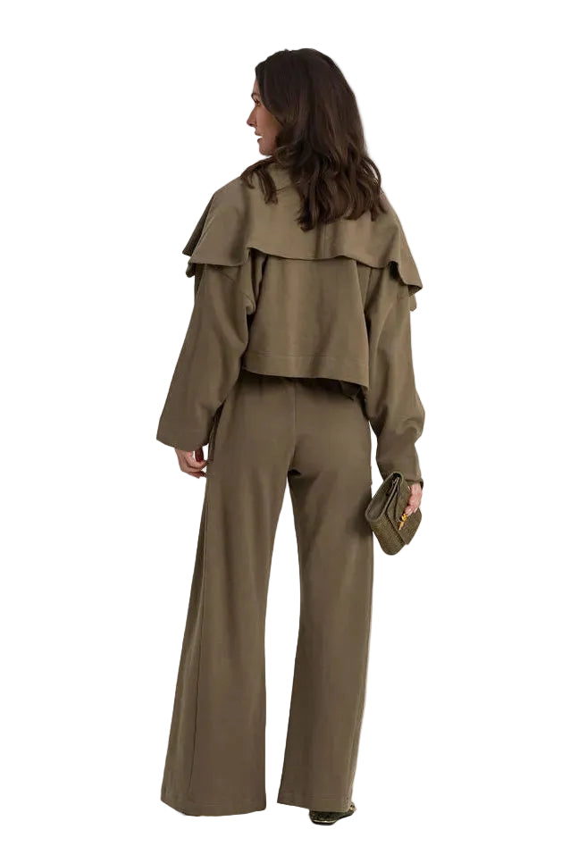 A model is shown from behind wearing a matching 100% cotton olive green two-piece set consisting of a ruffled long-sleeve crop top and wide-leg trousers, holding a small matching clutch against a plain white studio background.