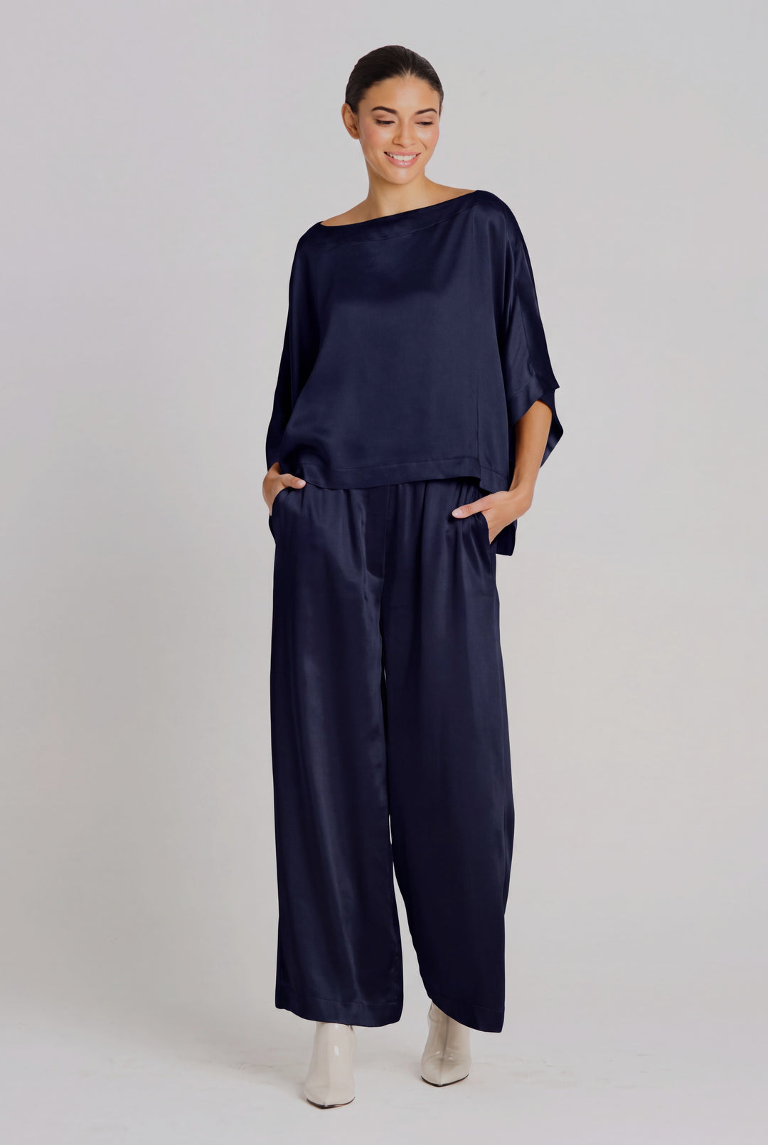 A woman modeling a navy midnight blue sateen two-piece set consisting of a wide-neck, elbow-length-sleeve top and matching wide-leg trousers, paired with white pointed-toe heels against a light gray studio background.