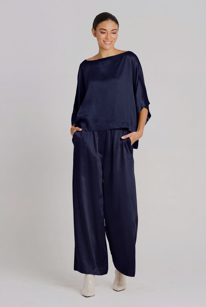 A woman modeling a navy midnight blue sateen two-piece set consisting of a wide-neck, elbow-length-sleeve top and matching wide-leg trousers, paired with white pointed-toe heels against a light gray studio background.