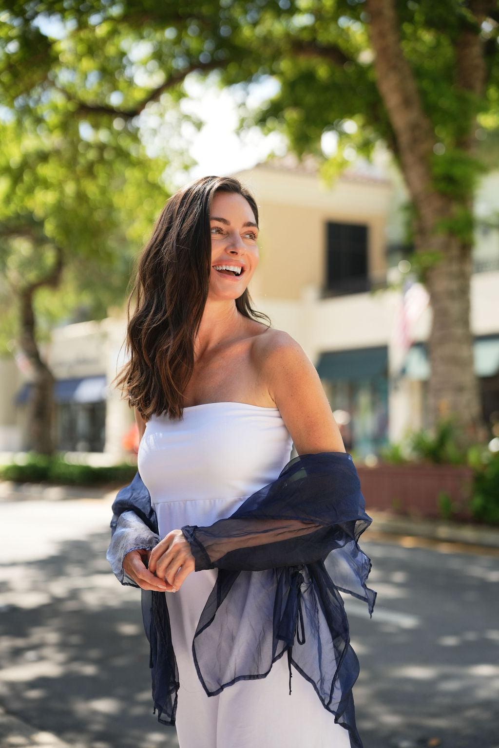 Woman in a white dress with an organza drawstring blue jacket standing outdoors with trees and buildings in the background.