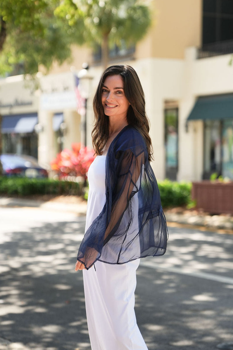 Woman in a blue organza top and white pants standing on a street with buildings and trees in the background