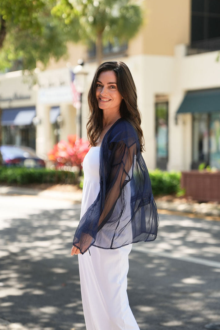 Woman in a blue organza top and white pants standing on a street with buildings and trees in the background