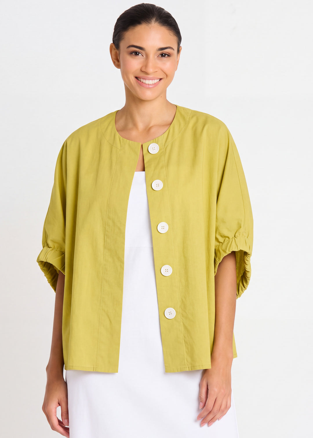 A woman modeling a short-sleeved chartreuse yellow nylon jacket with white buttons over a white dress like Mrs. Maisel, set against a plain white studio background.