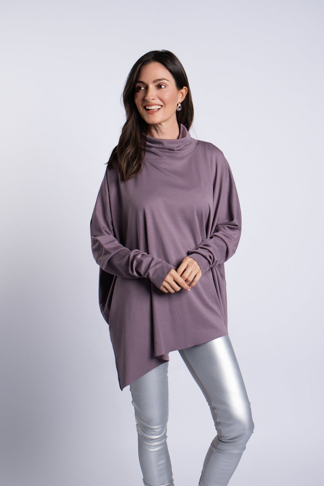 A woman modeling an oversized, long-sleeve cowl-neck pima cotton top in a muted mauve-purple color, paired with shiny metallic silver leggings and gray heels, against a light gray studio background.