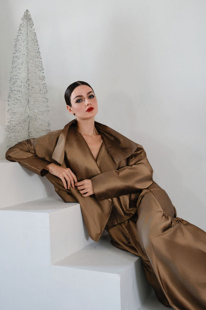 Woman in a brown dupioni silk outfit sitting on a white ledge with a white wall and decorative plant in the background.
