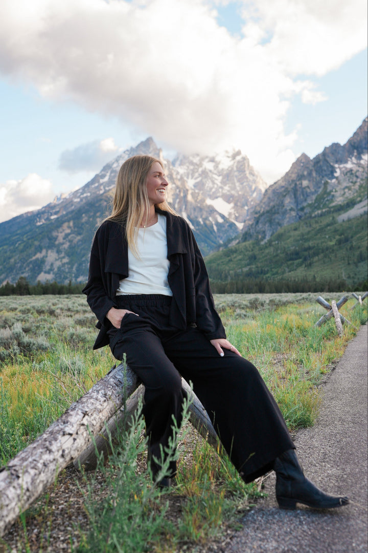 A woman wearing a black jacket, white top, black pants, and black boots sits on a log in a grassy field with mountains and a cloudy sky in the background.