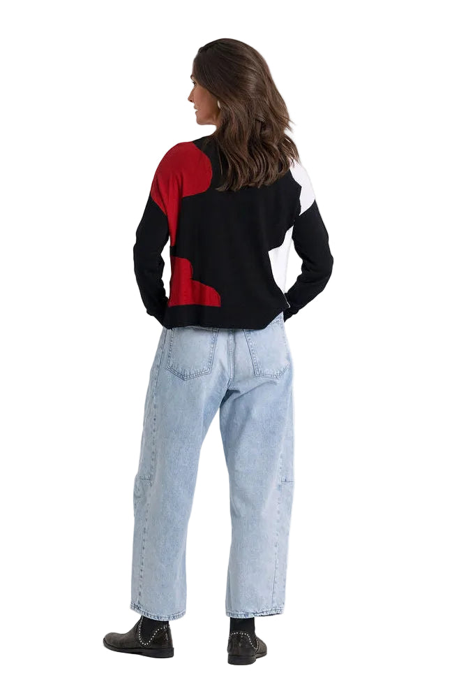 A woman viewed from the back wearing a 100% pima cotton color-block knit sweater with black, red, and white panels, paired with light-wash blue jeans and black shoes, against a plain white studio background.