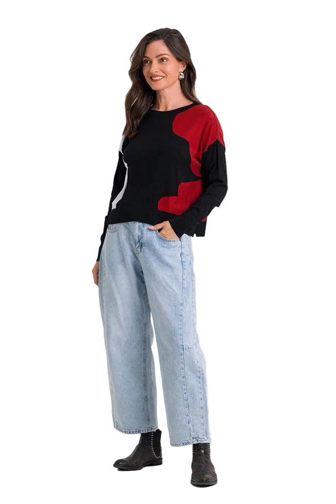 A woman modeling a 100% pima cotton color-blocked knit sweater with a black body and red shoulder/collar panels, paired with light-wash wide-leg jeans and dark ankle boots, against a plain white studio background.