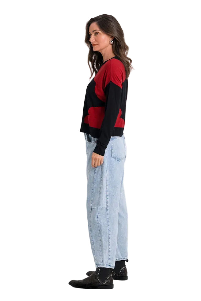 A woman stands in profile wearing a 100% pima cotton black and red striped knit sweater, light-wash blue jeans, and black shoes against a plain white studio background.