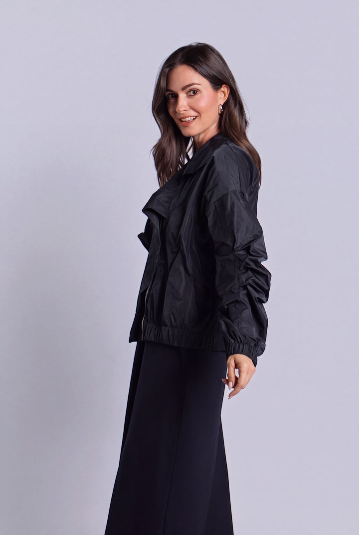 A woman modeling a black nylon lightweight jacket with gathered ruffle details on the shoulders and sleeves, paired with a long black skirt, against a solid light gray studio background.
