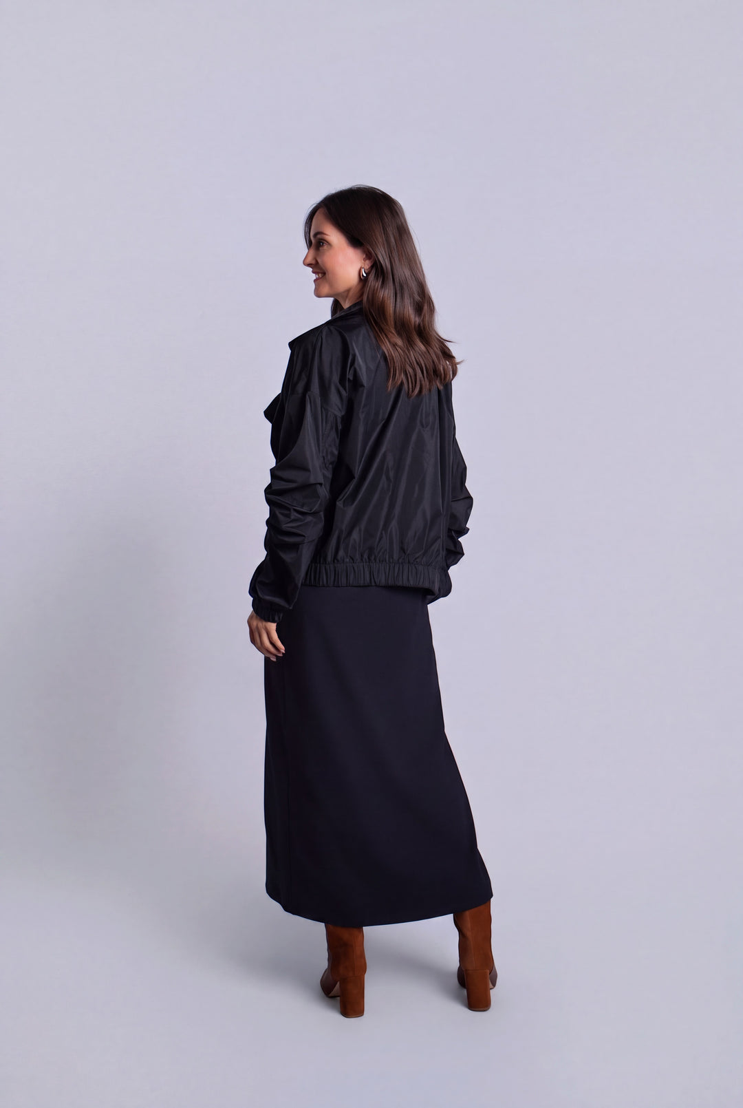 A woman is shown from the back wearing a black ruffled bomber jacket, a long black midi skirt, and brown boots, standing against a light gray studio background.