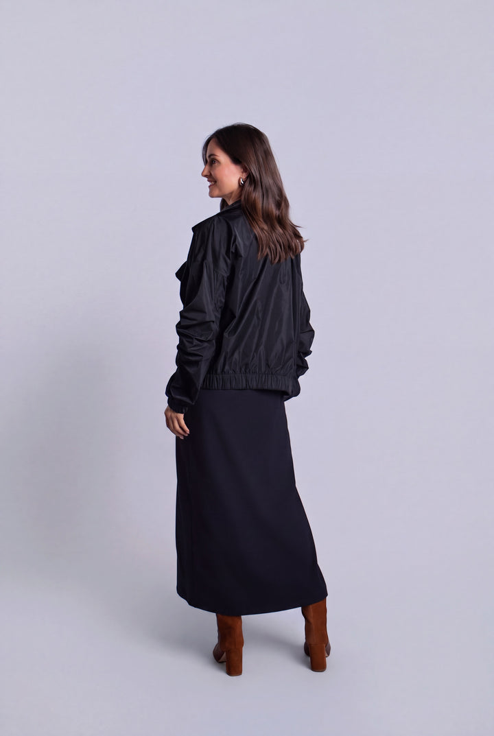 A woman is shown from the back wearing a black ruffled bomber jacket, a long black midi skirt, and brown boots, standing against a light gray studio background.