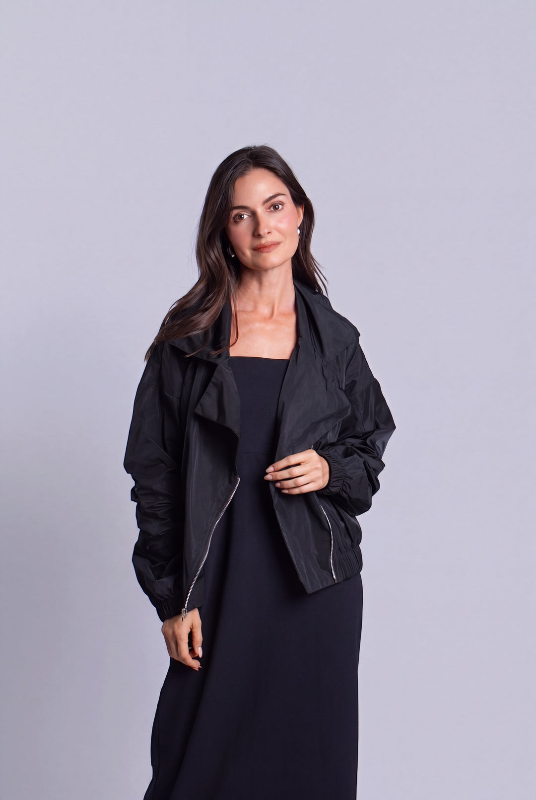 A woman modeling a black lightweight zip-front jacket over a black dress, standing against a plain light gray studio background.