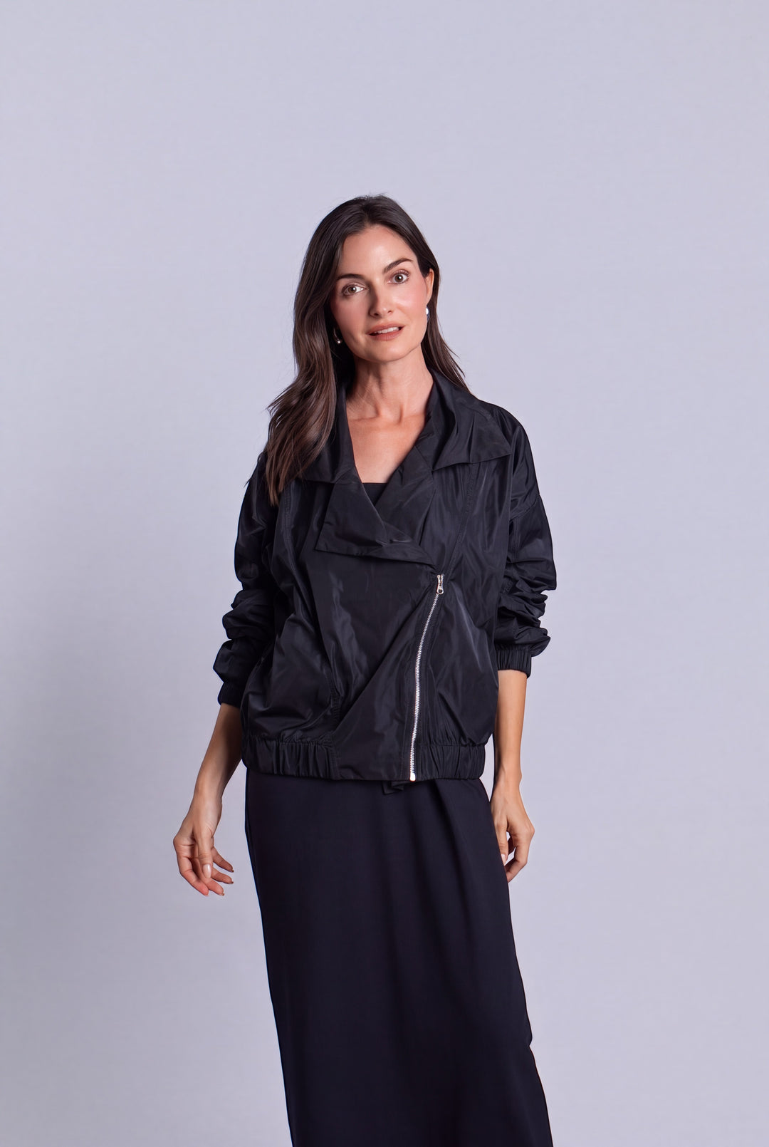 A woman modeling a black lightweight nylon jacket with a diagonal front zipper, paired with a long black skirt, against a solid light gray studio background.