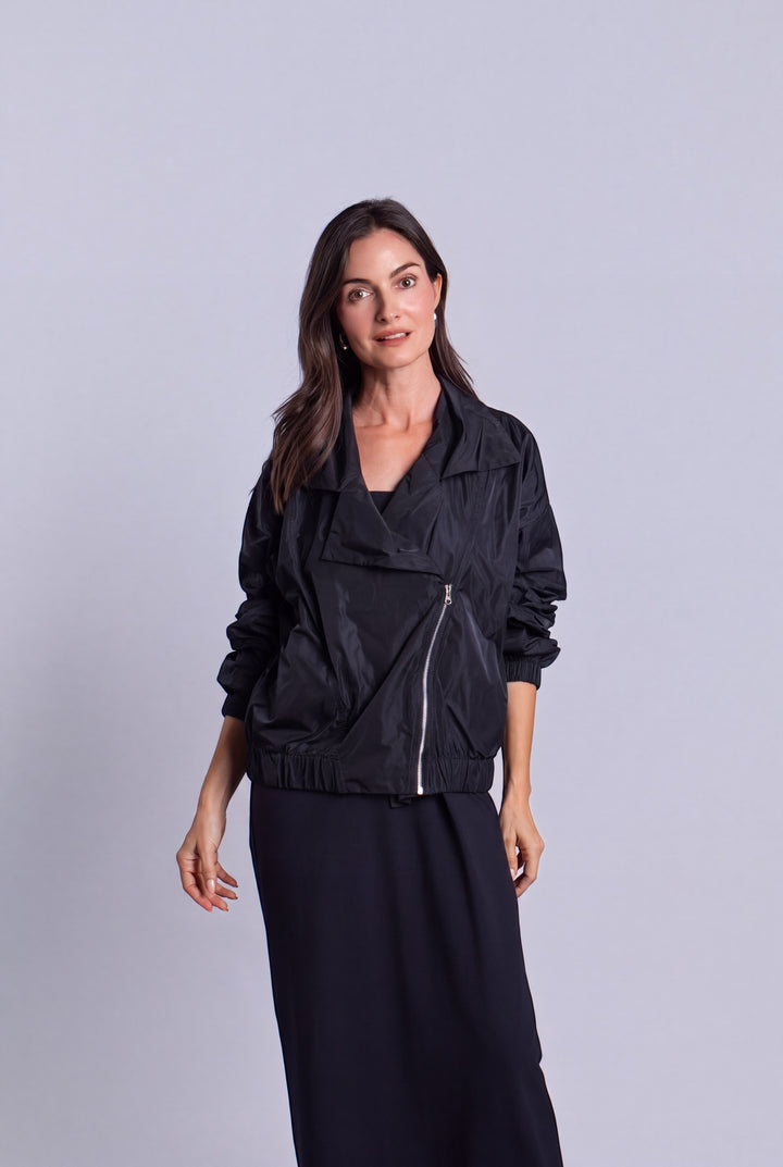 A woman modeling a black lightweight nylon jacket with a diagonal front zipper, paired with a long black skirt, against a solid light gray studio background.