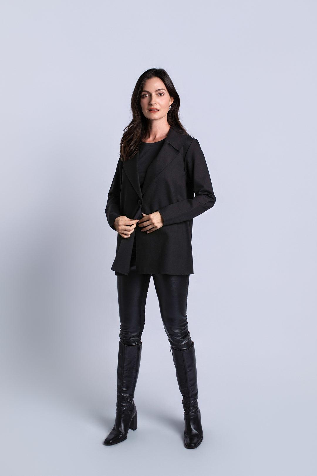 A woman modeling a black blazer, black top, black faux-leather pants, and black knee-high boots against a light gray studio background.
