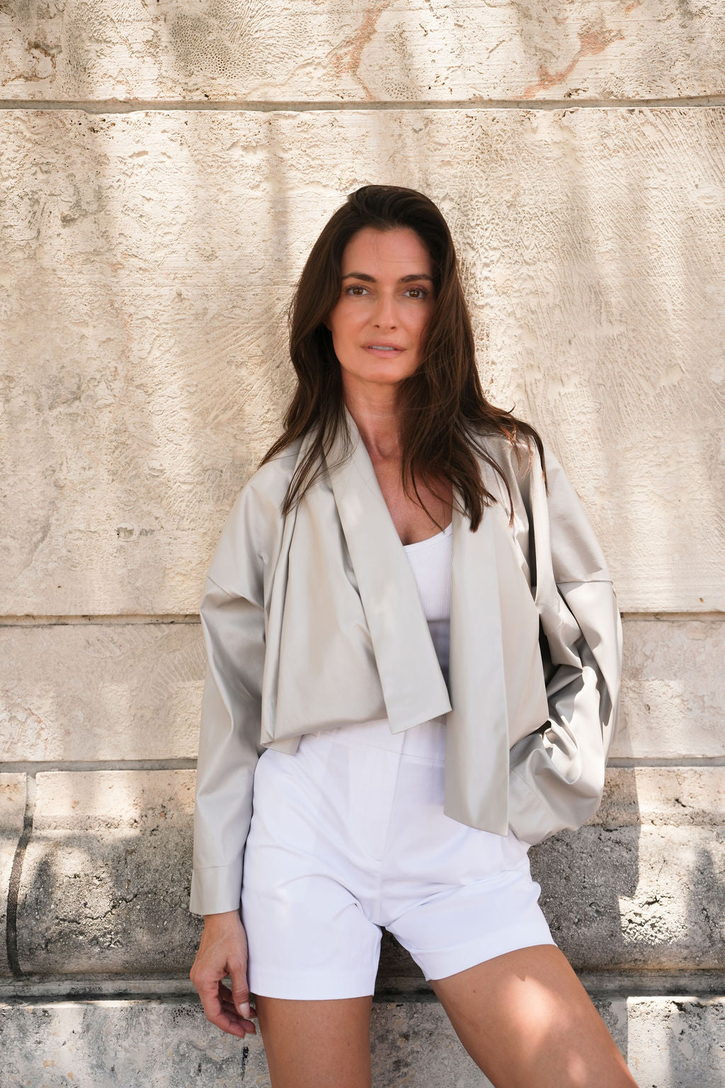 Woman in light gray jacket and white shorts standing against a textured stone wall in Coral Gables.