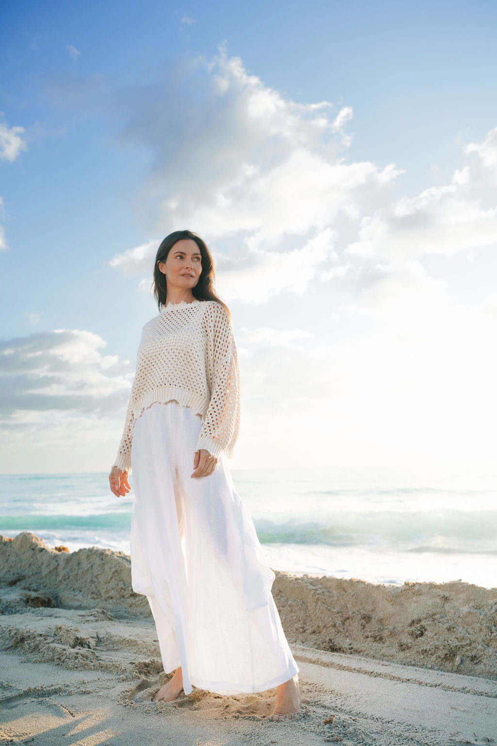 Woman standing on a beach wearing a beige knit sweater and white pants with a blue sky and clouds in the background.