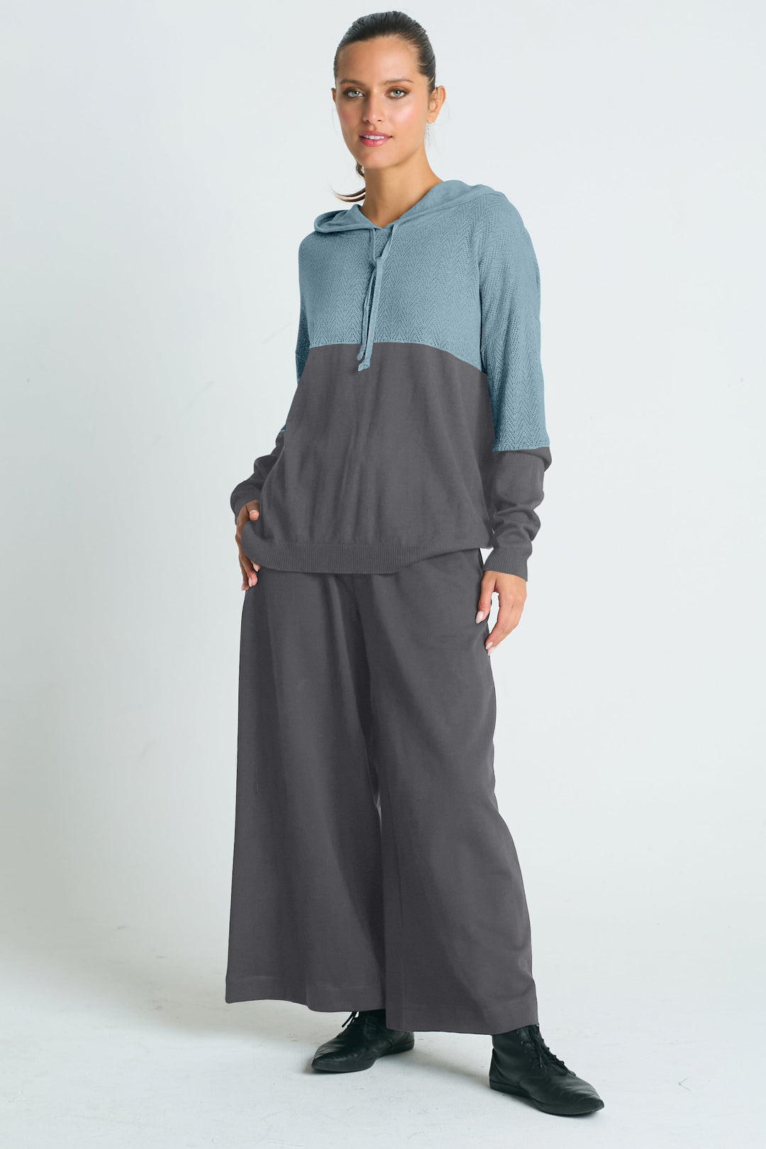 A model wearing a two-tone pointelle hoodie with a primarily black color on the lower half and a white color on the upper half, paired with black pants and black shoes.