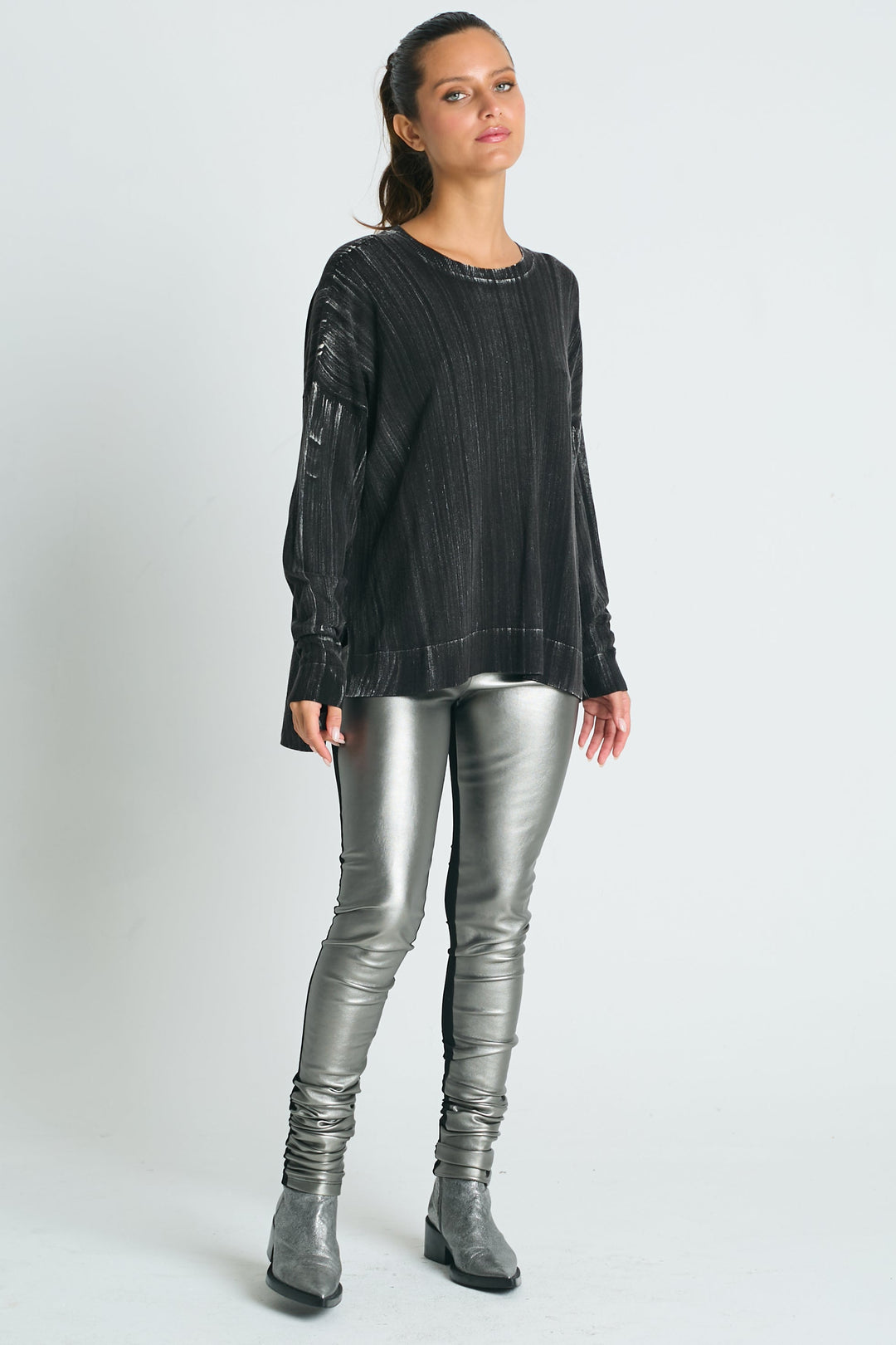 A female model wearing a long black crewneck sweater with metallic threads and silver leggings, standing against a white background.
