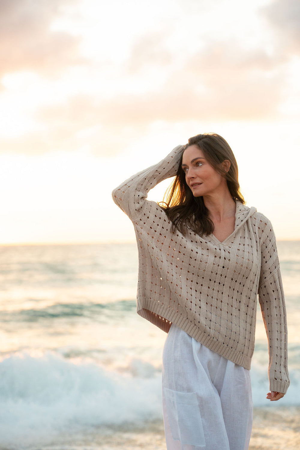 Woman in a beige patterned sweater and white pants standing on a beach at sunset.