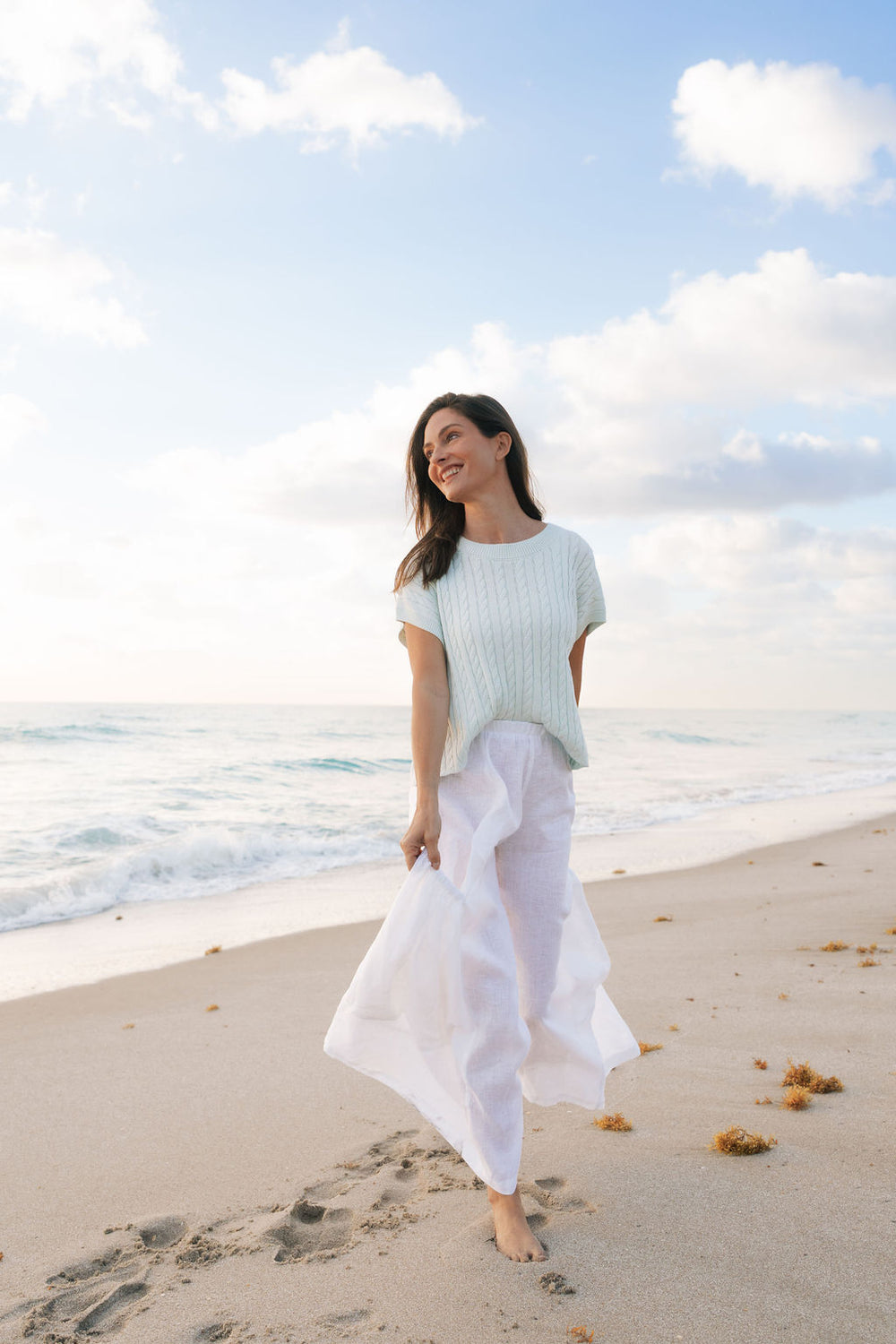 Woman walking on a beach with ocean and sky in the background in white pants