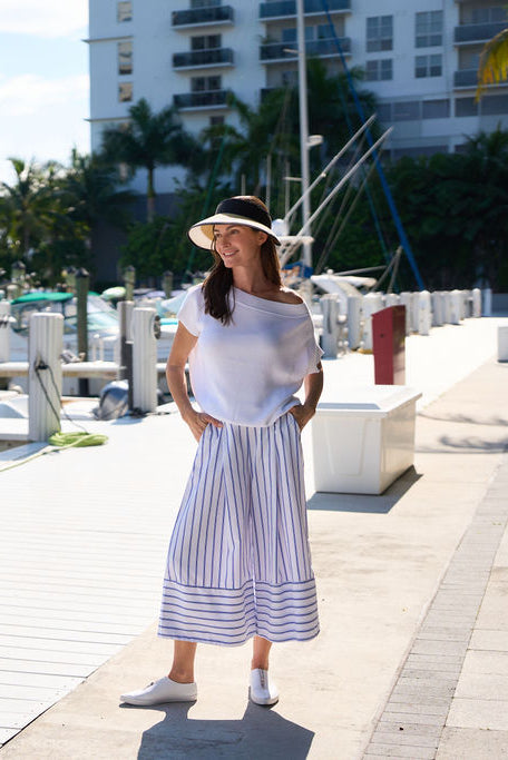 Woman in a white pima cotton knit top and striped pants standing on a dock with buildings and palm trees in the background.