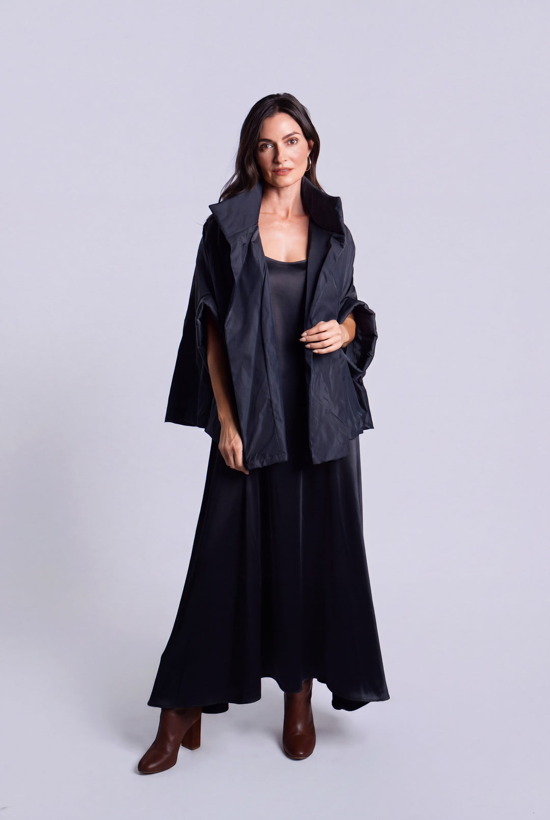 A woman modeling a dark navy blue outfit consisting of a draped cape-style jacket, a sleeveless top, wide-leg skirt, and brown ankle boots, shot against a solid light gray studio background.