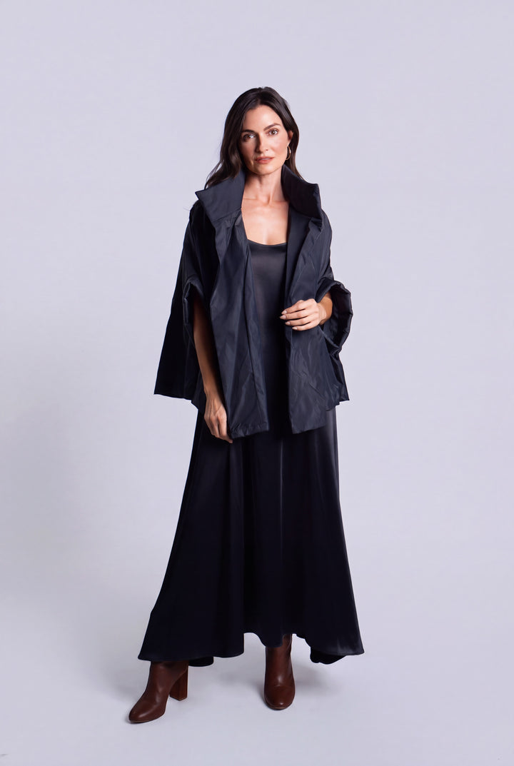A woman modeling a dark navy blue outfit consisting of a draped cape-style jacket, a sleeveless top, wide-leg skirt, and brown ankle boots, shot against a solid light gray studio background.
