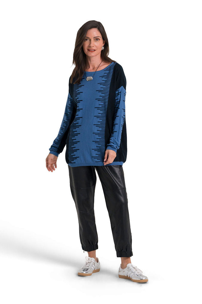 A woman modeling a long-sleeve pima cotton top with a blue and black ruffled design, paired with black pants and white sneakers, against a plain white background.