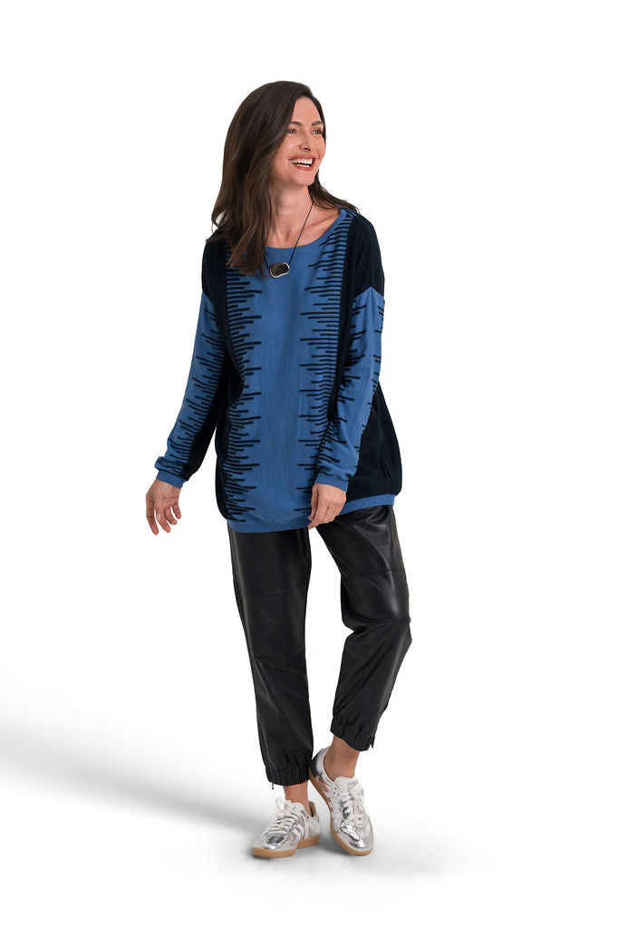 A woman modeling a long-sleeve tunic top with a textured blue and black design, paired with black pants and white sneakers, set against a plain white background.
