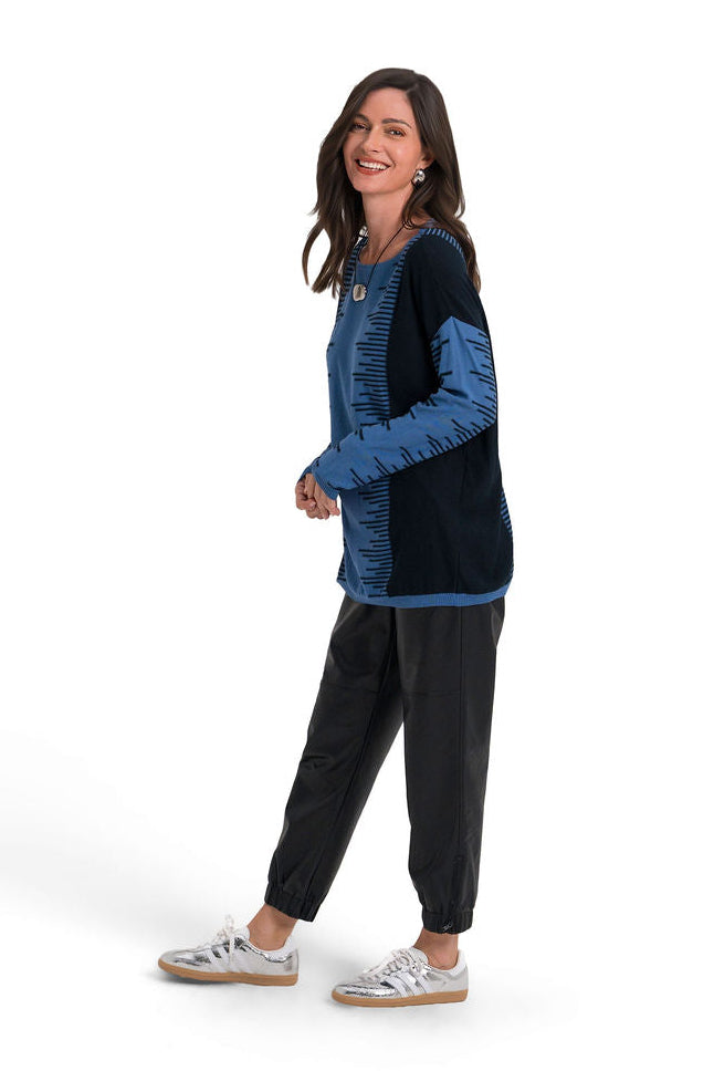 A woman modeling a long-sleeve pima cotton knit top with a color-blocked design in blue and black, paired with black pants and white sneakers, set against a plain white background.