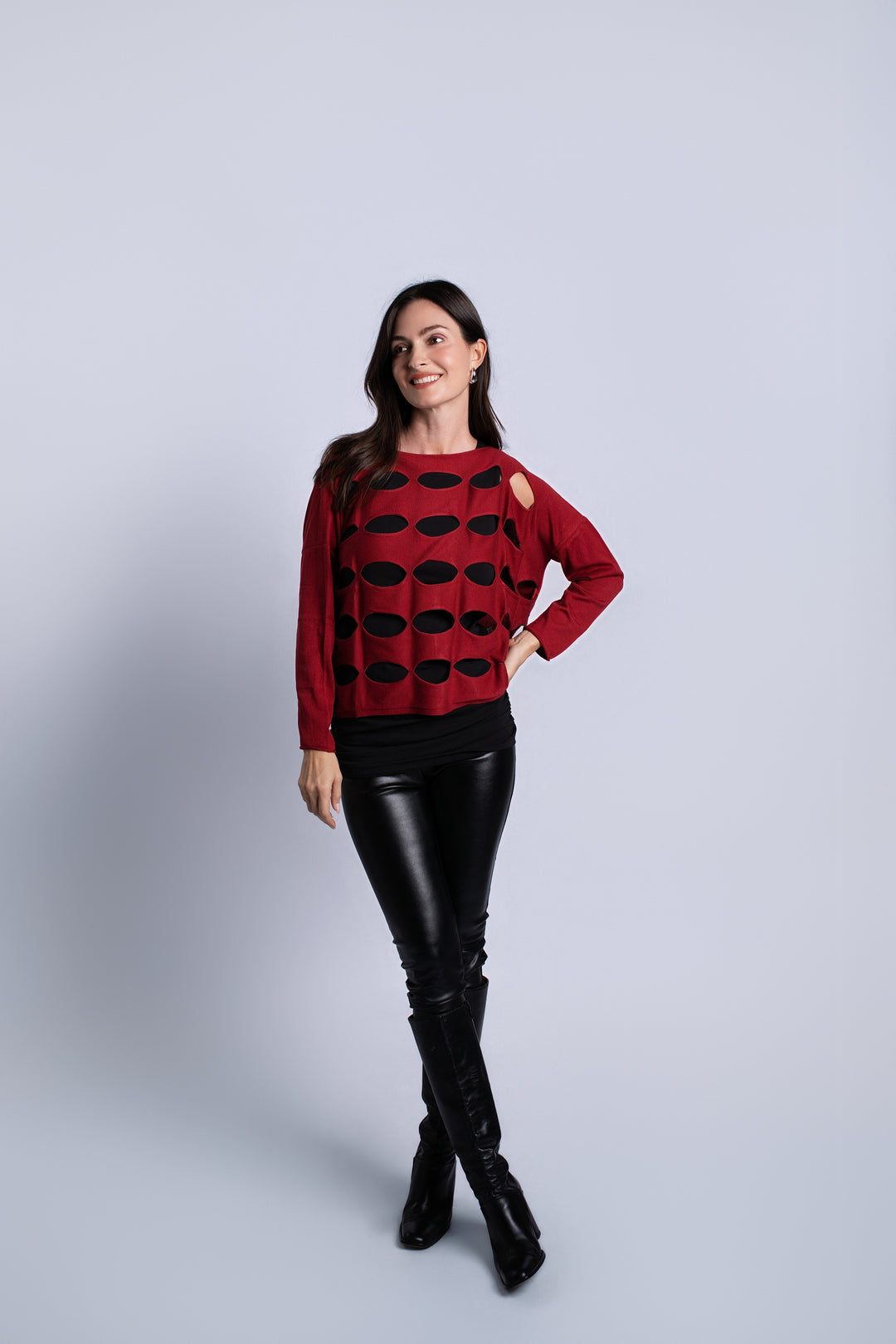 A woman modeling a red knit sweater with black circular cutout patterns, paired with black faux-leather pants and black knee-high boots, against a light gray studio background.