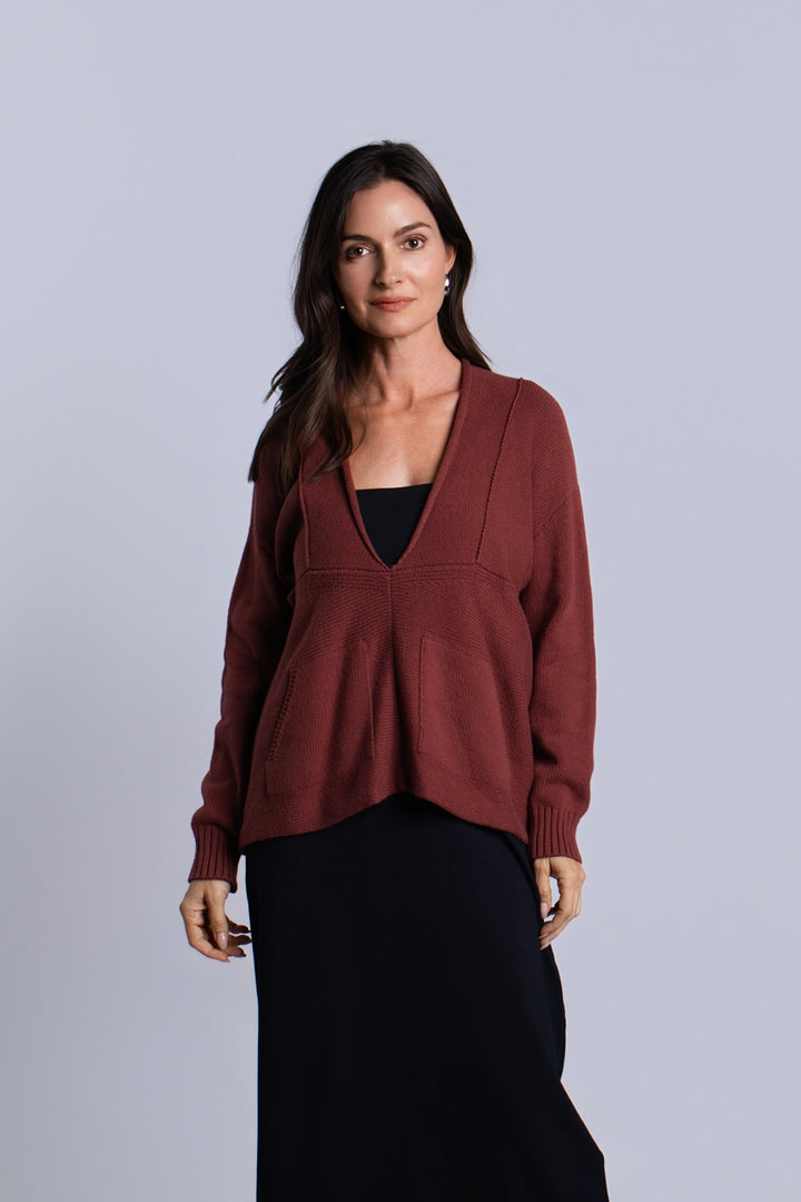 A woman modeling a rust-brown pima cotton knit cardigan with a split V-neckline and front pockets, worn over a black top and black skirt against a light gray studio background.