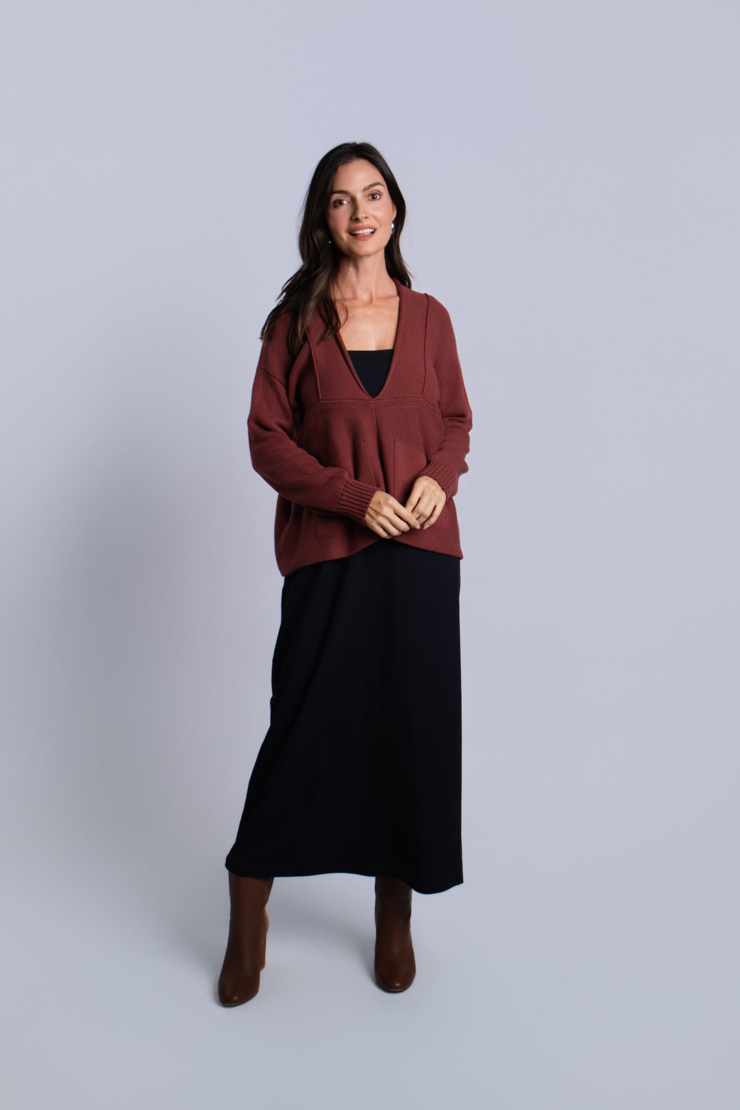 A woman modeling a rust-brown pima cotton cardigan sweater over a black top and black midi skirt, paired with brown ankle boots, against a solid light gray studio background.