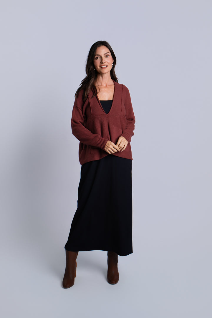 A woman modeling a rust-brown pima cotton cardigan sweater over a black top and black midi skirt, paired with brown ankle boots, against a solid light gray studio background.