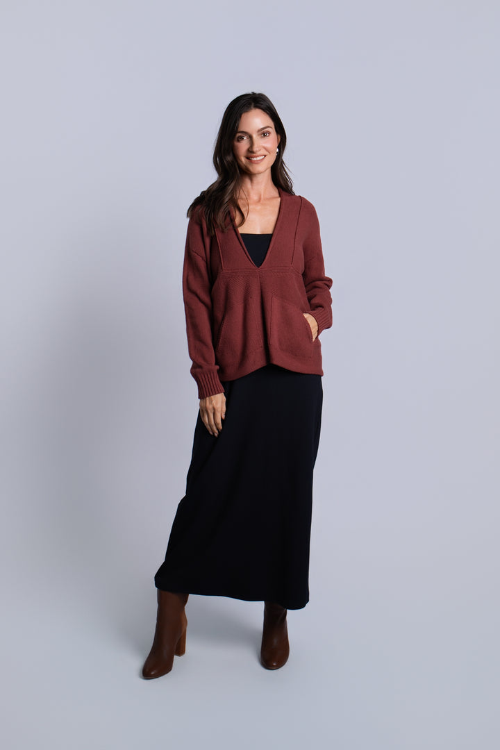 A woman modeling a rust-brown knit pima cotton cardigan sweater over a black top and black midi skirt, paired with brown ankle boots, against a light gray studio background.