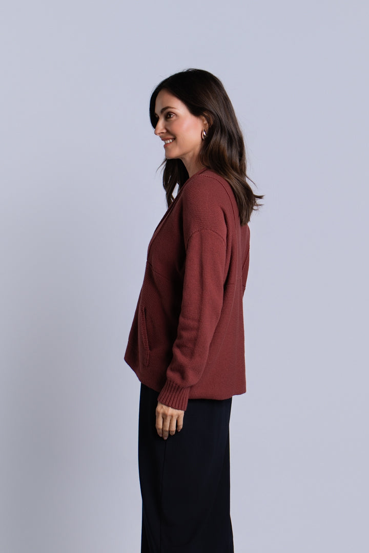 A woman in side profile wearing a rust-brown long-sleeve knit sweater and dark navy pants, standing against a solid light gray studio background.