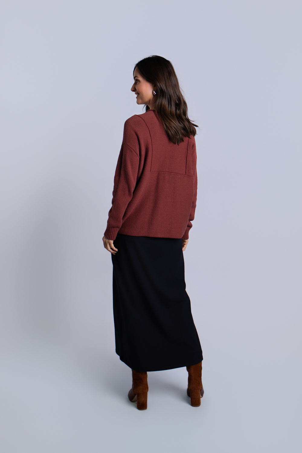 A woman is shown from the back wearing a rust-brown long-sleeve pima cotton knit top and a black midi skirt, paired with brown ankle boots, against a light gray studio background.
