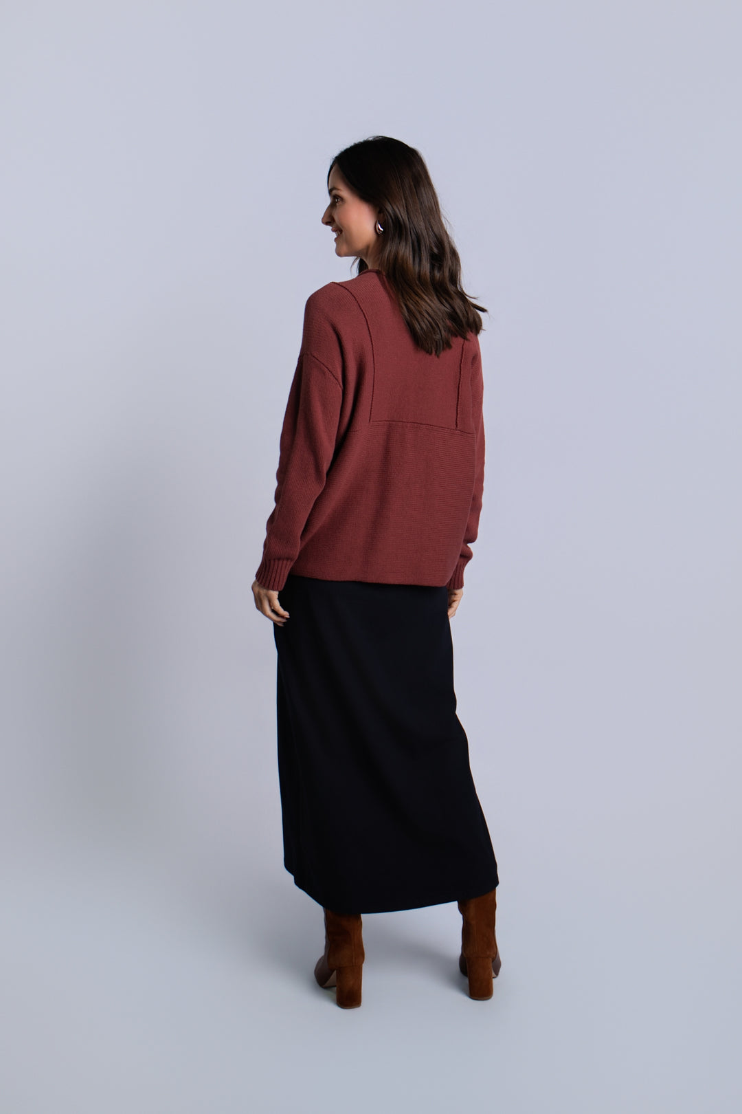 A woman is shown from the back wearing a rust-brown long-sleeve pima cotton knit top and a black midi skirt, paired with brown ankle boots, against a light gray studio background.