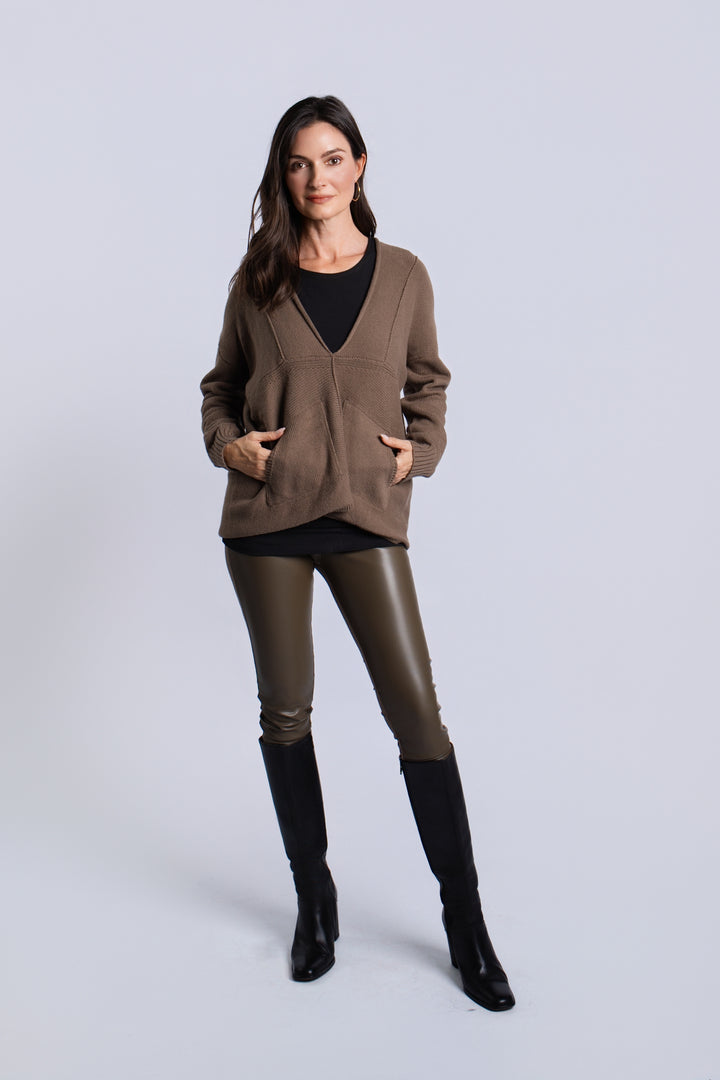 A woman modeling a brown open-front cardigan with pockets over a black top, paired with dark brown faux-leather pants and black knee-high boots, against a light gray studio background.