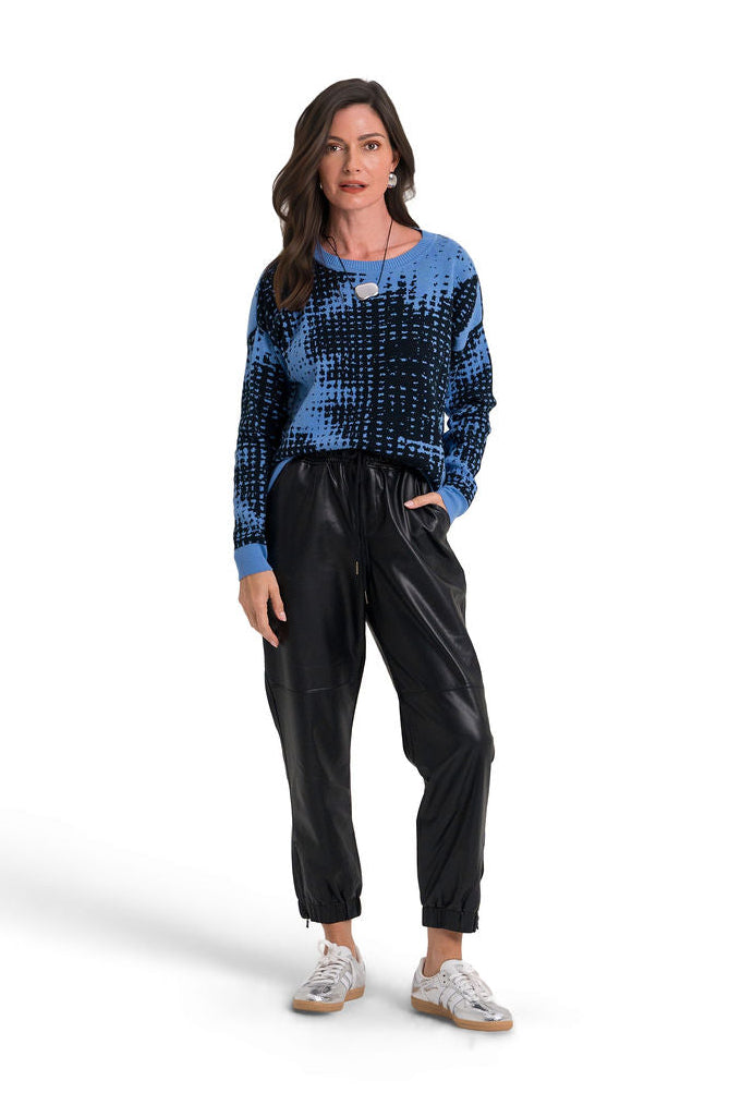 A woman modeling a blue and black patterned pima cotton knit sweater paired with black pants and silver sneakers, standing against a plain white studio background.
