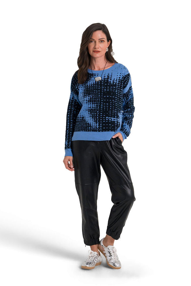 A woman modeling a blue and black patterned pima cotton knit sweater, black pants, and white sneakers against a plain white background.